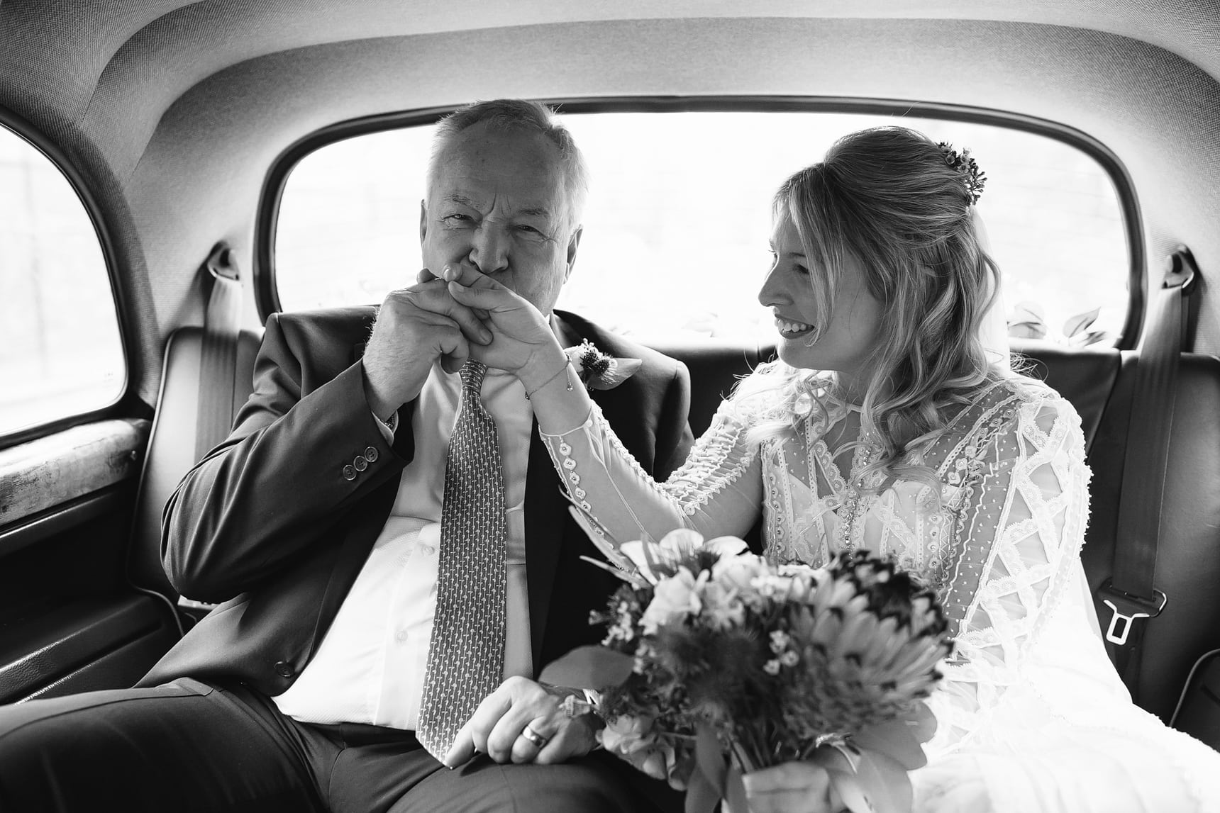 Black and white photo in a pack of a wedding car with father kissing his daughters hand