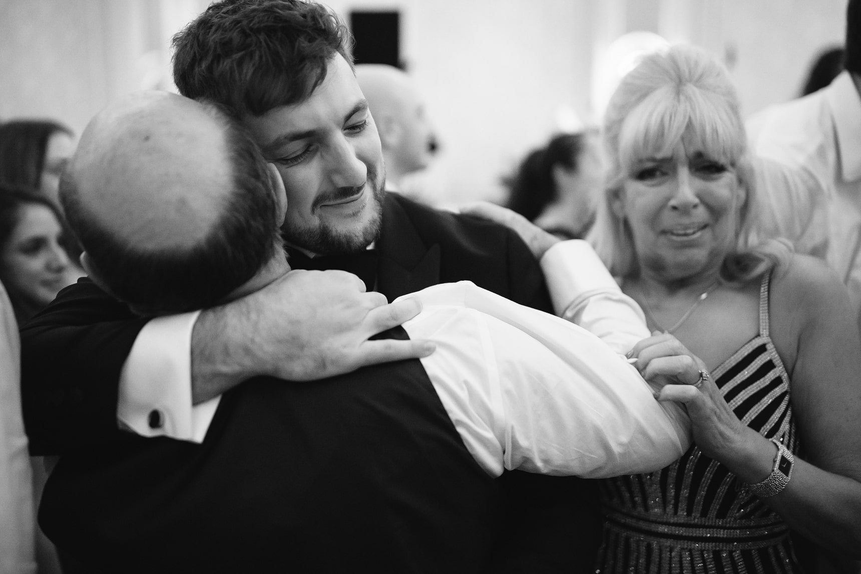 Black-and-white photograph of a groom hugging his father with his mother in the background